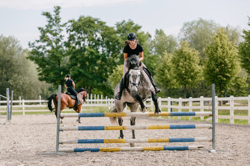 Girl on a dapple gray horse practicing jumping over a log fence in the drill hall of the equestrian...