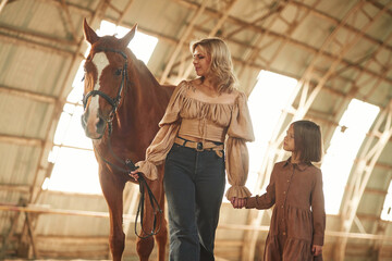 Woman with her little daughter is with horse indoors