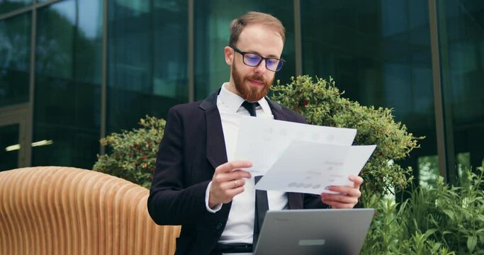 Businessman reading documents while sitting on bench outdoor. Man working on laptop remotely from office wearing suit cheking document doing job. Freelance worker, project on the go outdoor.