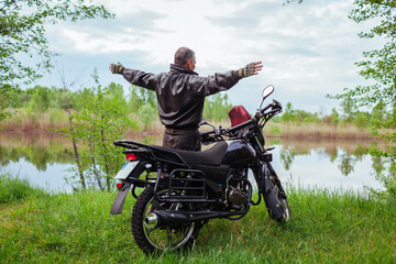 Back view of happy senior biker raising arms enjoying river landscape after riding motobike....