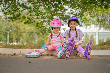 Two sisters in protective sports uniforms, helmet and knee pads are sitting resting after roller...