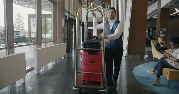 Hotel bellboy wearing uniform is walking along corridor pushing cart with luggage working alone. Porter occupation and guest service concept.