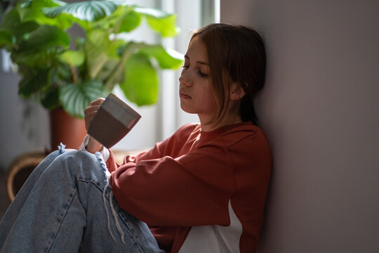 Frustrated sad teenage girl sitting on floor with cup devastated thinking about trouble, broken heart. Young woman have bad mood due to hormones in period. Depressive thoughts from problems at school.