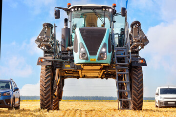 A tractor sprayer with a high wheelbase stands next to cars in a field against a blue cloudy sky.