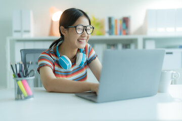 A young Girl or student using a laptop computer in the library while wearing glasses and headphones. She is smiling and looking happy.