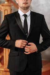 Portrait of an adult man buttoning up a black jacket in his room. Preparation of the groom for the wedding ceremony.