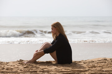 Beautiful young blonde woman in black shirt sits on the shore of the beach sad and depressed. The woman is pensive and looks at the ground exhausted and tired. The woman is suffering.