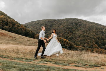 A romantic fairytale couple of newlyweds are running in a field at sunset, behind high mountains covered with trees. The bride in a white wedding dress, the groom in a white shirt and black pants