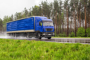 A blue truck with a semi-trailer transports cargo against the backdrop of a forest in rainy weather along a slippery highway, industry
