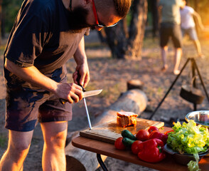 A man with a beard and black glasses sharpens a knife in nature while cooking. Outdoor recreation in summer. Bright and fresh vegetables on the table, bushcraft