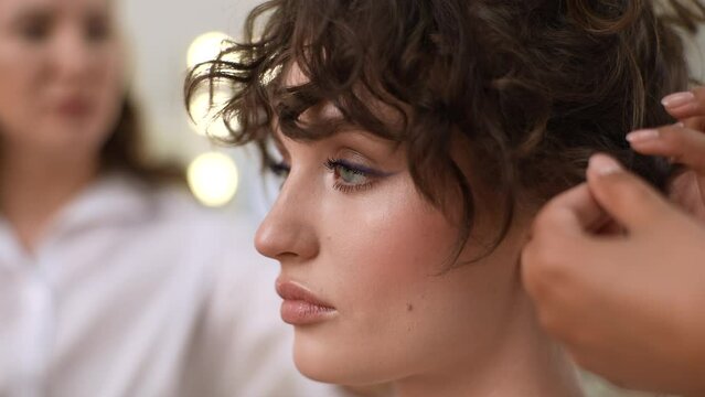 Close-up face of elegance young woman being given beautiful model hairstyle in modern beauty studio. Hairdresser making final touch to lady long curly hair. Hairdresser giving beauty salon service.