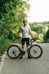 Obraz premium Photo of a sporty male cyclist in gear posing for the camera with a professional road bike outside the city on a road and forest background.