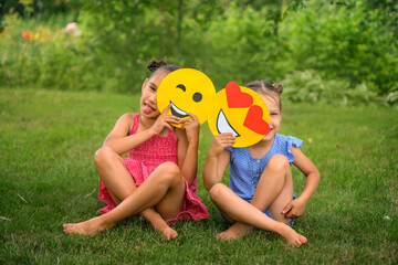 Playful laughing funny girls hide behind a variety of smile faces sitting in the park on a lawn on a sunny summer day