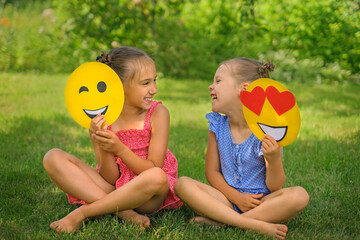 Playful funny girls laugh looking at each other, holding emoticons with hearts and a winking funny smile face