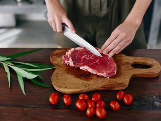 Woman with knife in hand cutting fresh steak meat for roasting in kitchen with salt pepper and...