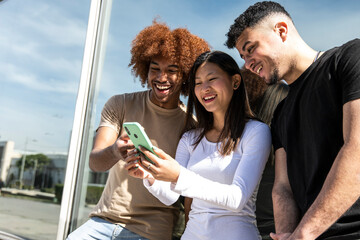 Three young diverse people looking at a smartphone standing int the street.Two young men smiling and watching at the phone of a happy woman.