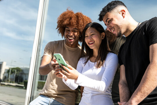 Three Young Diverse People Looking At A Smartphone Standing Int The Street.Two Young Men Smiling And Watching At The Phone Of A Happy Woman.