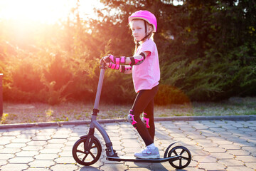 A beautiful girl in a pink protective outfit rides a scooter through the streets of the city in summer. Safe cycling and scootering without injury. © HENADZY