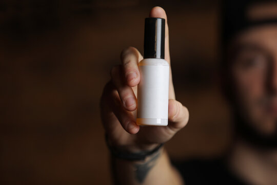 Unfocus Young Man Holding In Hand White Plastic Bottle With Mock Up For Text On Dark Background. Focus On Packaging Bottle. Grooming For Skin, Hair Men, Beauty Concept. Spa Salon, Barbershop