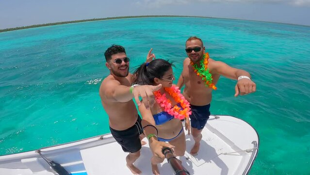 Selfie of a small company of two young latin men and woman wearing swimsuits dancing on the bow of a yacht. Turquoise water of the sea and a tropical beach with palm trees on the background