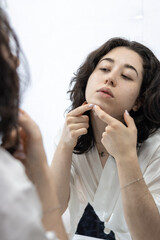 Unhappy young latin lady in white bathrobe standing in front of mirror in bathroom, removing blackheads from her face skin. Acne, pimples, problem skin care concept