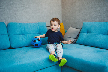 A two-year-old little boy sits on the sofa with a ball in his hands. A developed emotional boy plays with a ball at home