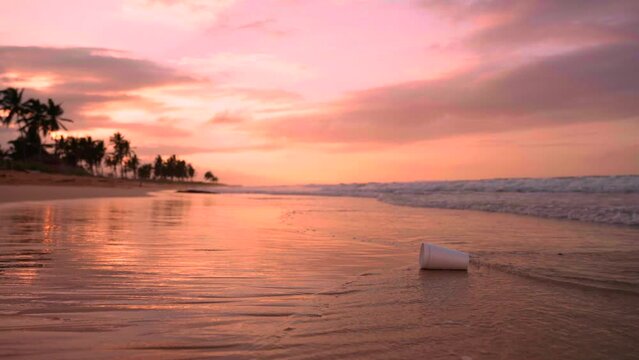 White foam cup on the beach floating on the waves at beautiful sunset. People leave plastic waste and trash on the seashore. Environment protection of nature