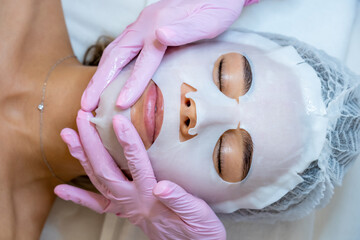 Professional doctor cosmetologist applying sheet mask on face of customer, woman in aesthetic clinic