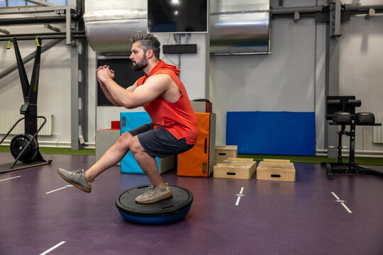 Fit athletic man performing exercise on gymnastic hemisphere bosu ball in gym.