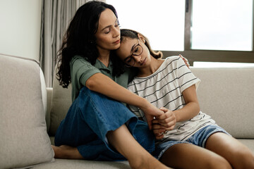 Caring mother comforting her teenage daughter, both sitting on the couch at home, sharing a tender...