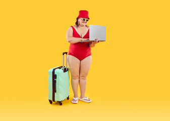 Happy funny fat woman in red hat and swimsuit with suitcase getting ready for holiday trip booking tickets online via laptop isolated on studio yellow background. Vacation and summer journey concept.