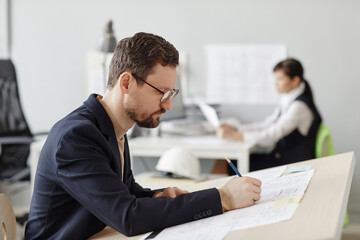 Side view portrait of male engineer at drawing blueprints and plans at table in office, copy space