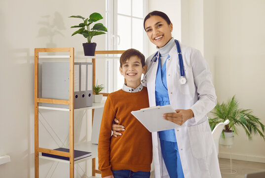 Portrait Of A Friendly Smiling Female Doctor Pediatrician Holding Report File With Appointment And Hugging A Child Boy Patient During Medical Examination In Clinic Or At The Hospital.