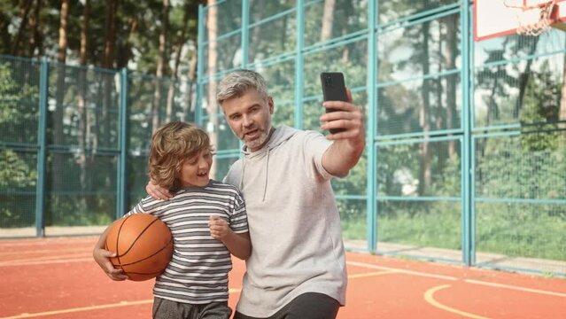 Smiling man takes photo on mobile phone with his child. Mature father with little son taking selfie picture with smartphone on background of basketball court. Bright family memories