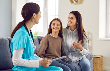 Family doctor talks to mother and child during home visit. Happy, smiling mother having conversation with woman pediatrician. Friendly paediatrician, mum and daughter sitting on sofa and talking