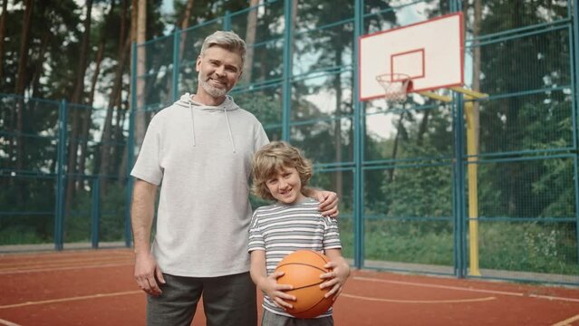 Portrait of caring father and son smiling, posing and looking at camera on background of basketball court. Family spending time together. Dad and teenage child standing together on playground.