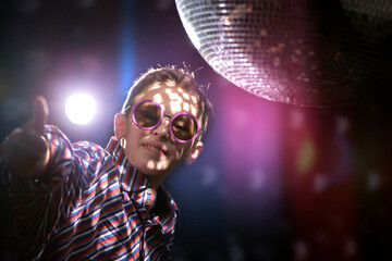 A young boy in stylish glasses shows a class sign against the background of light rays from a disco ball. Children's disco, party.