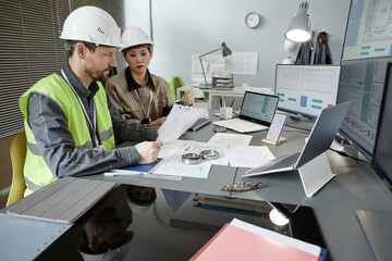 Wide angle view at two engineers designing blueprints at workplace in office and wearing hardhats, copy space