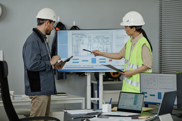 Side view portrait of two engineers reviewing blueprints indoors and pointing at computer screen,...