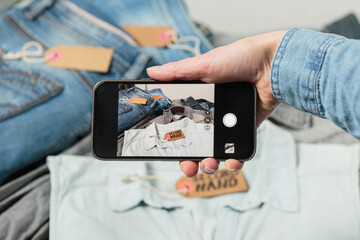 View of womans hand taking photo of denim clothes to upload in second hand fashion app on the smartphone. Sell, buy and swap clothes online