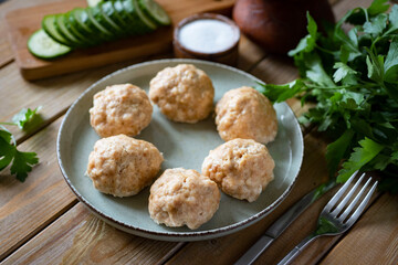 A dietary dish for the whole family: steamed meat meatballs in a beautiful plate, next to a bunch of fresh herbs and sliced cucumbers on wooden boards. A dish for children: pork meatballs. Close-up