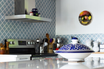Blue and white painted dish with cover set on quartz counter, with blue tiled kitchen in soft focus background