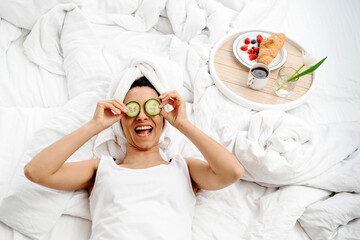 A young beautiful woman just got out of the shower lay down on a snow-white bed and put slices of cucumbers to her eyes. On the bed is a tray with a wonderful breakfast, a cup of coffee and a flower.