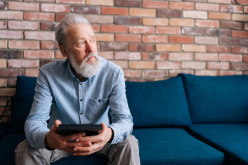 Front view of sad bearded gray-haired mature adult male sitting on couch at home holding picture of wife in frame and crying, looking away. Concept of nostalgia, grief, longing, loneliness in old age.