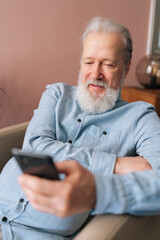 Vertical portrait of happy mature male holding smartphone using mobile online app, smiling looking to screen sitting on armchair. Bearded older man enjoying social media at home after retirement.