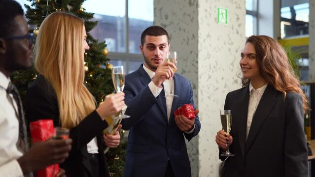 Multiethnic corporate workers celebrate Christmas party clinking and toasting glasses next to Christmas tree. Happy diverse people drinking champagne and holding New Year presents in an office.
