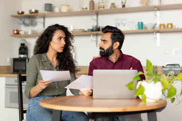 Worried arab spouses sitting at desk in front of laptop, checking bills, working on family budget in kitchen interior