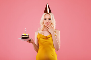 Happy pleased girl in party hat holding piece of cake and closing mouth with hand, celebrating...