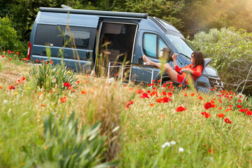 Van life theme: A dark-haired woman in a red dress sits in an armchair, glass in hand, relaxing in a meadow of blooming red poppies in front of a camper van.  Travelling Greece, wanderlust life style. © Martin Mecnarowski