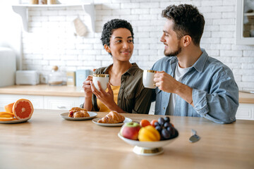 Happy couple in love of different nationalities, sit und hugging at home in the kitchen, dressed in stylish clothes, drinking morning coffee with croissants, look at each other, smile